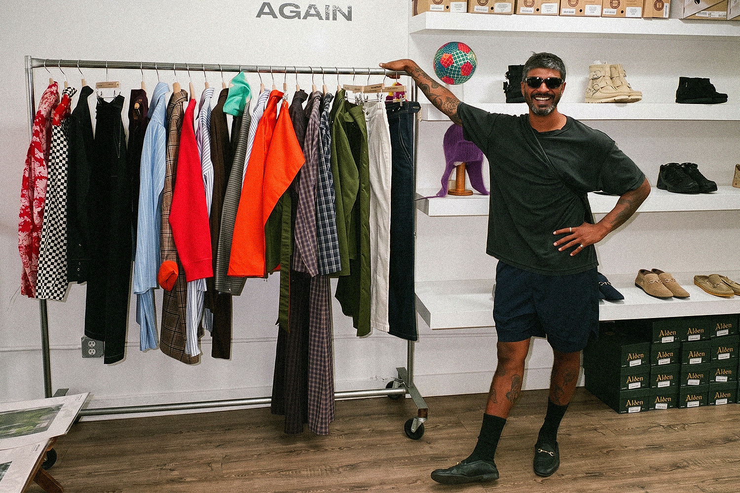Mayan Rajendran in sunglasses and a dark T-shirt stands next to a clothing rack in a bright store with shelves of shoes