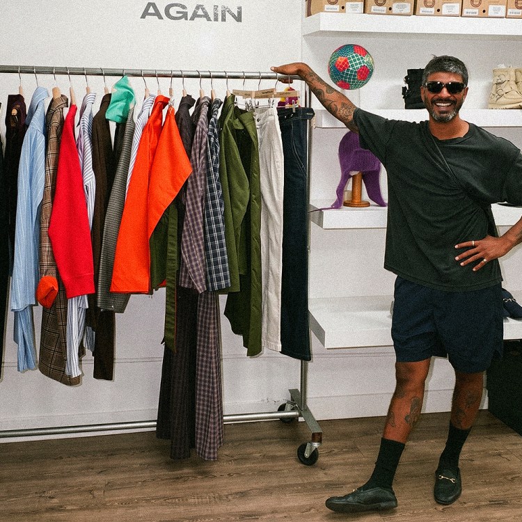 Mayan Rajendran in sunglasses and a dark T-shirt stands next to a clothing rack in a bright store with shelves of shoes