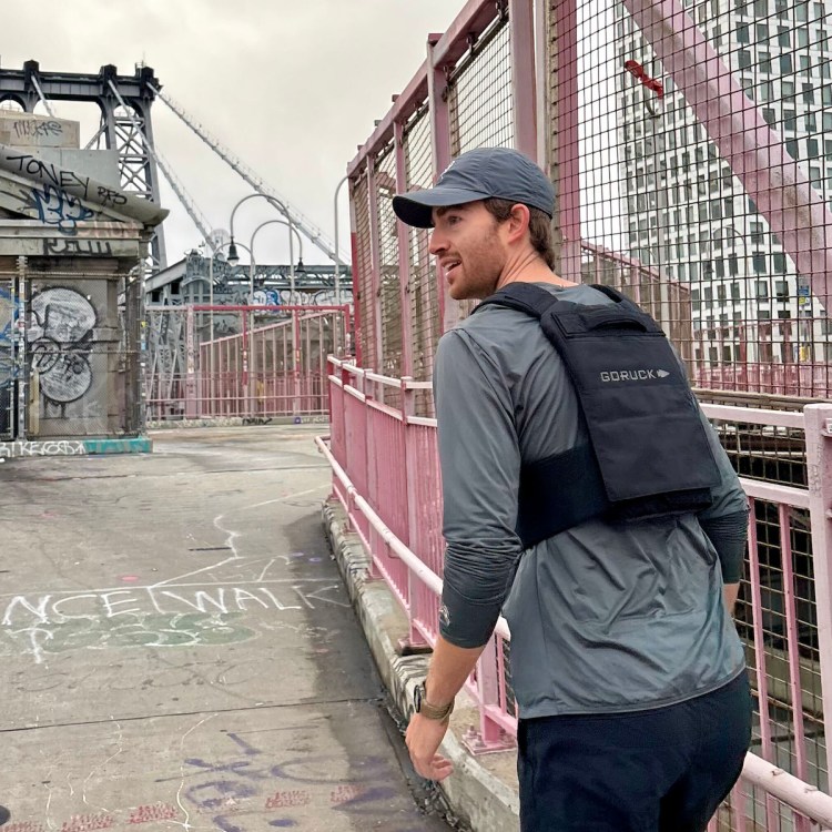 Man in a gray shirt and black GORUCK vest walks on a graffiti-covered bridge with pink railings.