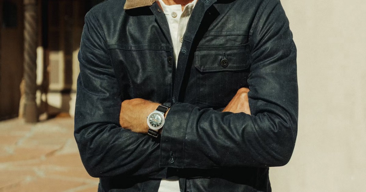 Man in a dark blue jacket with a tan corduroy collar, white henley, and a watch on his crossed arms.
