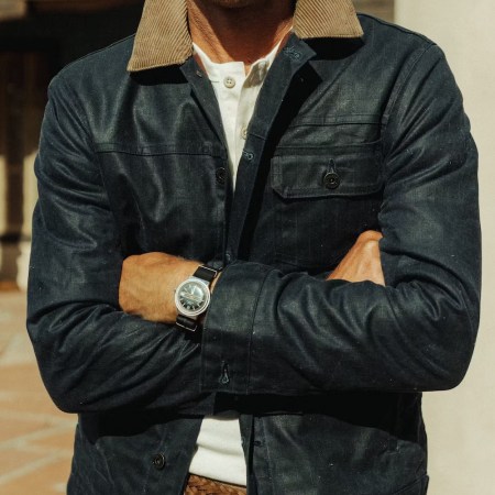 Man in a dark blue jacket with a tan corduroy collar, white henley, and a watch on his crossed arms.