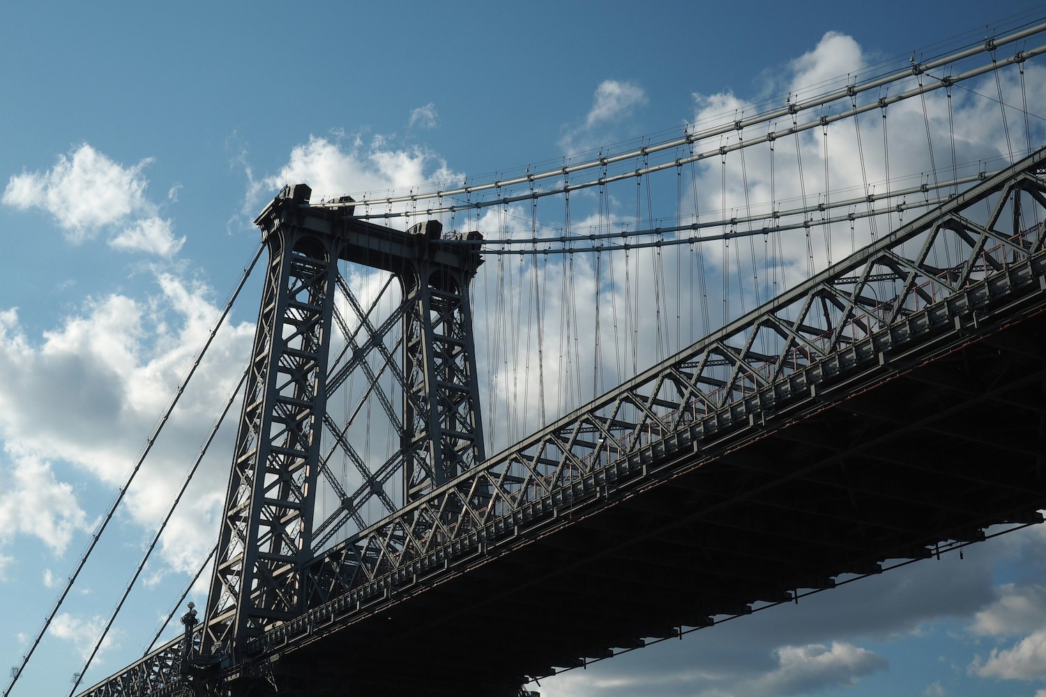 The Williamsburg Bridge's dark steel structure and suspension cables against a blue sky with white clouds.
