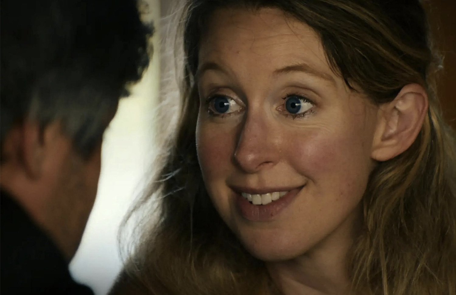 Elizabeth Holmes, A fair-skinned woman with blue eyes and long blonde hair smiles, looking toward a man whose back is visible.
