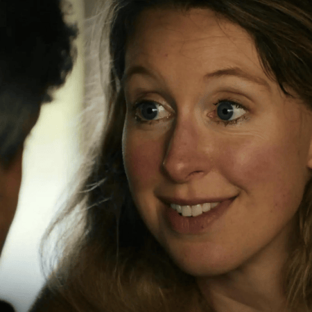 Elizabeth Holmes, A fair-skinned woman with blue eyes and long blonde hair smiles, looking toward a man whose back is visible.