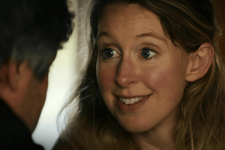 Elizabeth Holmes, A fair-skinned woman with blue eyes and long blonde hair smiles, looking toward a man whose back is visible.